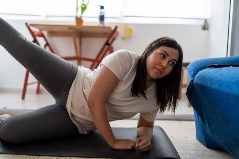 Mulher de aproximadamente 50 anos realizando agachamento com o peso do próprio corpo na sala de estar, com luz natural ao fundo.